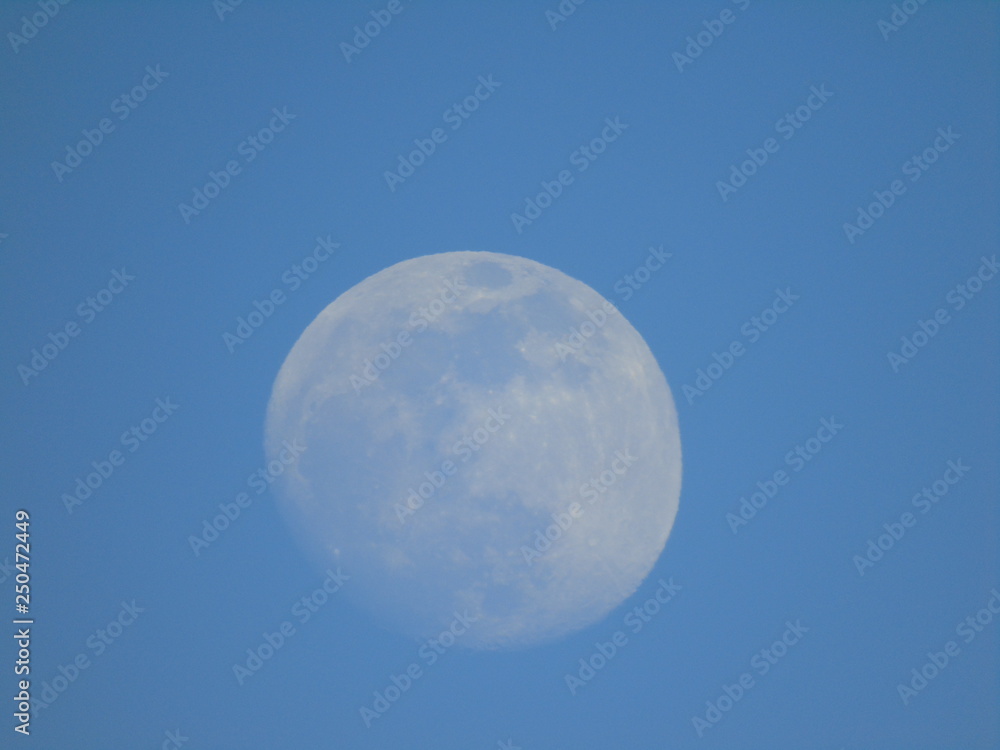 Liguria, Italy - 02/18/2019: An amazing photography of the full moonlight over the mountains, hidden and out of the trees in the village by day with beautiful blue sky in the background in winter days Liguria, Italy - 02/18/2019: An amazing photography of the full moonlight over the mountains, hidden and out of the trees in the village by day with beautiful blue sky in the background in winter days