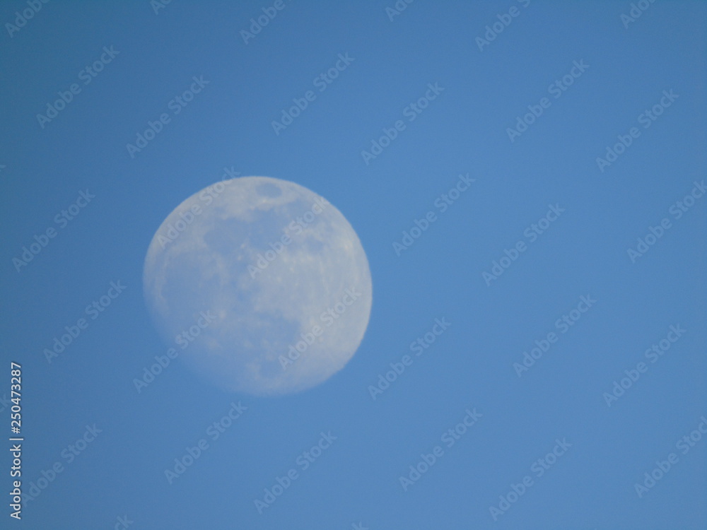 Liguria, Italy - 02/18/2019: An amazing photography of the full moonlight over the mountains, hidden and out of the trees in the village by day with beautiful blue sky in the background in winter days Liguria, Italy - 02/18/2019: An amazing photography of the full moonlight over the mountains, hidden and out of the trees in the village by day with beautiful blue sky in the background in winter days