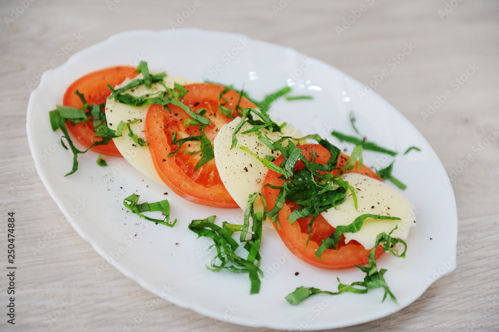 Caprese salad on a white plate. Large slices of tomatoes, mozzarella cheese and basil greens Caprese salad on a white plate. Large slices of tomatoes, mozzarella cheese and basil greens
