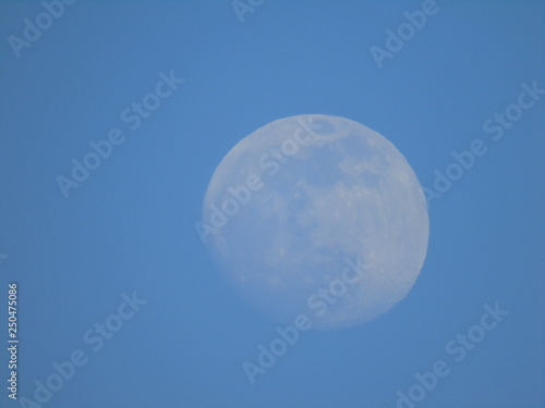 Liguria, Italy - 02/18/2019: An amazing photography of the full moonlight over the mountains, hidden and out of the trees in the village by day with beautiful blue sky in the background in winter days