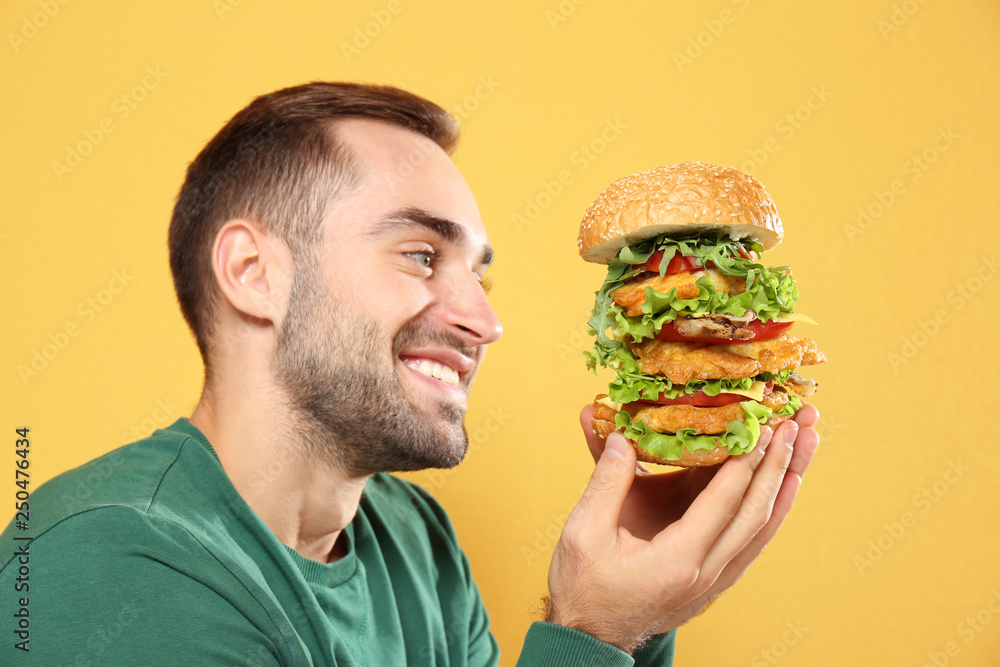 Young hungry man eating huge burger on color background Young hungry man eating huge burger on color background