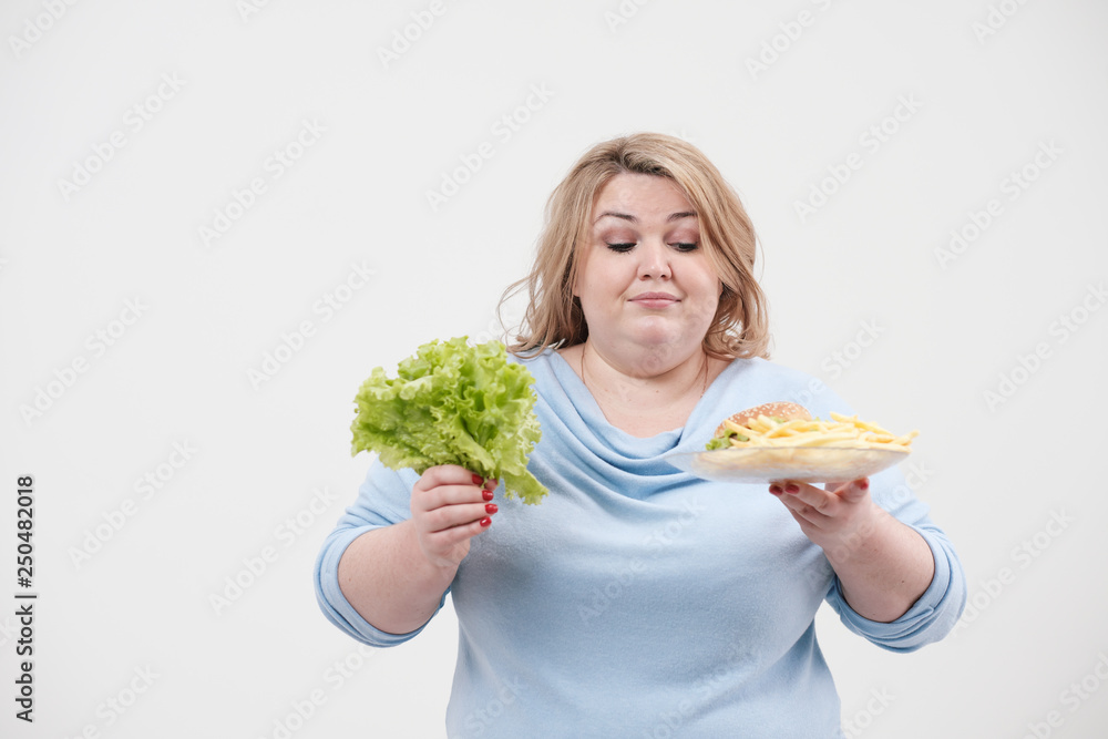 Young fluffy fat woman in casual blue clothes on a white background holding green salad leaves and a plate of fast food, hamburger and fries. Diet and proper nutrition. Young fluffy fat woman in casual blue clothes on a white background holding green salad leaves and a plate of fast food, hamburger and fries. Diet and proper nutrition.