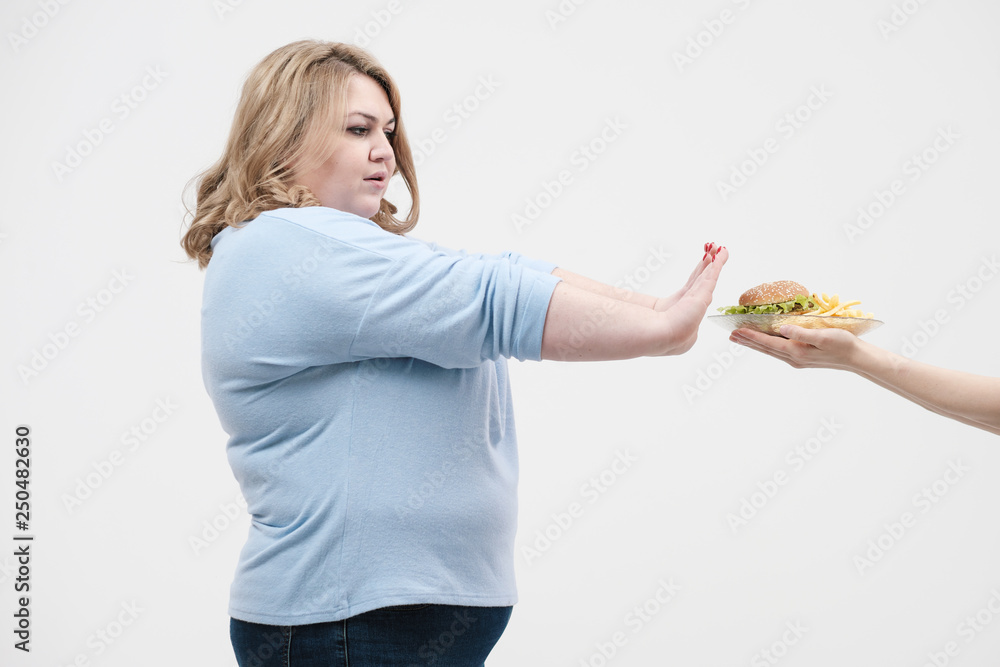 A young magnificent fat woman in casual blue clothes on a white background refuses from the fast food offered to her, a hamburger and french fries. Diet and proper nutrition. A young magnificent fat woman in casual blue clothes on a white background refuses from the fast food offered to her, a hamburger and french fries. Diet and proper nutrition.