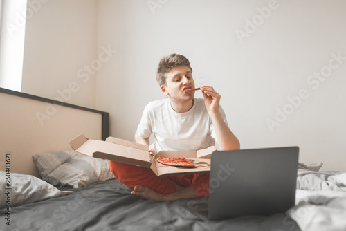 Young man sits on a bed with a pizza box and a laptop. Student sitting on the bed eats a pizza and uses a laptop. Rest houses with internet, movie and pizza
