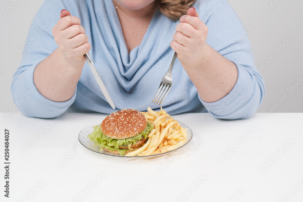Young curvy fat woman in casual blue clothes on a white background at the table eagerly eating fast food, hamburger and french fries. Diet and proper nutrition. Young curvy fat woman in casual blue clothes on a white background at the table eagerly eating fast food, hamburger and french fries. Diet and proper nutrition.