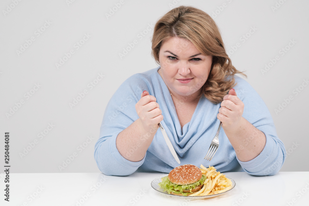 Young curvy fat woman in casual blue clothes on a white background at the table eagerly eating fast food, hamburger and french fries. Diet and proper nutrition.