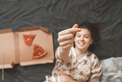 Portrait of a young girl wearing a pajamas pajamas, lying in a bed with a box of pizza and showing a heart symbol with fingers. Girl loves pizza. Weekend concept.