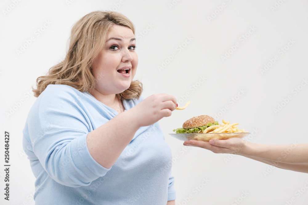 Young curvy fat woman in casual blue clothes on a white background sneaking off a plate of hamburger and french fries. Diet and proper nutrition. Young curvy fat woman in casual blue clothes on a white background sneaking off a plate of hamburger and french fries. Diet and proper nutrition.
