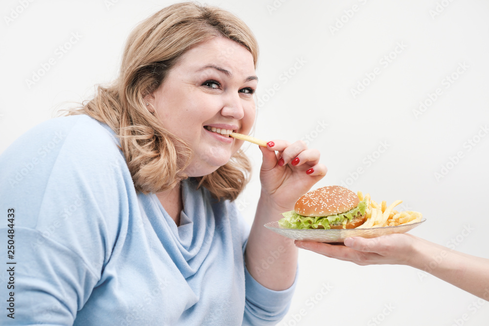 Young curvy fat woman in casual blue clothes on a white background sneaking off a plate of hamburger and french fries. Diet and proper nutrition. Young curvy fat woman in casual blue clothes on a white background sneaking off a plate of hamburger and french fries. Diet and proper nutrition.