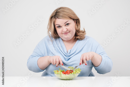 Young lush fat woman in casual blue clothes on a white background at the table and eats a vegetable salad with tomatoes. Diet and proper nutrition.