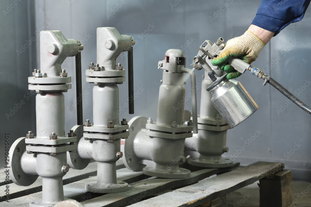 A worker at a factory in a special room paints parts of the valves from the spray gun. Paint valves in gray color, hand and spray gun close up A worker at a factory in a special room paints parts of the valves from the spray gun. Paint valves in gray color, hand and spray gun close up