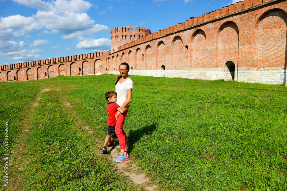 Son hugs mother in warm summer day under blue sky with clouds and Smolensk medieval city red brick wall with tower on the background, Russia