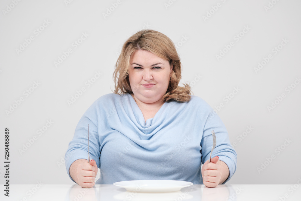 Young curvy fat woman in casual blue clothes on a white background at the table sitting in front of an empty plate and starving. Diet and proper nutrition. Young curvy fat woman in casual blue clothes on a white background at the table sitting in front of an empty plate and starving. Diet and proper nutrition.