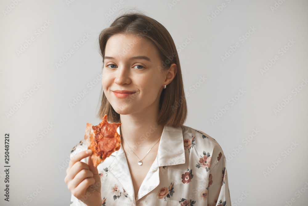 Funny girl in a pajama with a piece of pizza in her hands looks at the camera and smiles with a smirk. Cute girl eating pizza on a light background. Isolated.