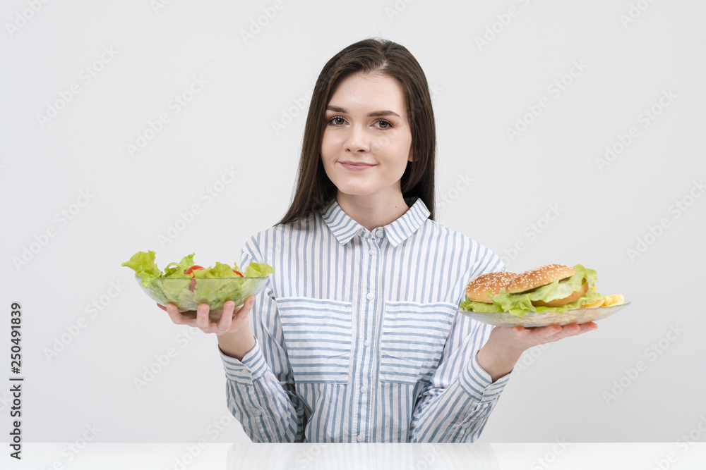 Slender brunette girl on a white background chooses between a plate of fast food and hamburgers and healthy food salad. Slender brunette girl on a white background chooses between a plate of fast food and hamburgers and healthy food salad.