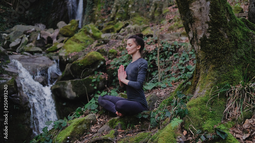 Young slim woman practicing yoga outdoors in moss forest on background of waterfall. Unity with nature concept. Girl meditates sitting