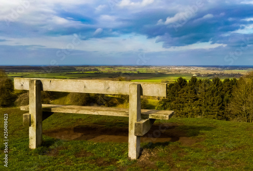 Landscape of an unoccupied wooden park bench on the top of a hill overlooking the Warwickshire countryside. Spring sunshine with blue skies and clouds. Burton Dassett, England.