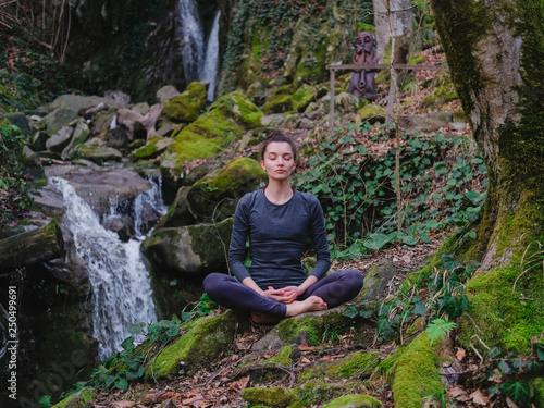 Young slim woman practicing yoga outdoors in moss forest on background of waterfall. Unity with nature concept. Girl meditates sitting