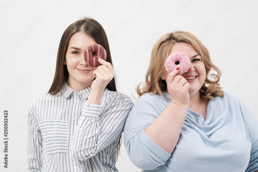 Two girls, thin and fat on a white background, are holding pink glazed donuts in their hands. Two girls, thin and fat on a white background, are holding pink glazed donuts in their hands.
