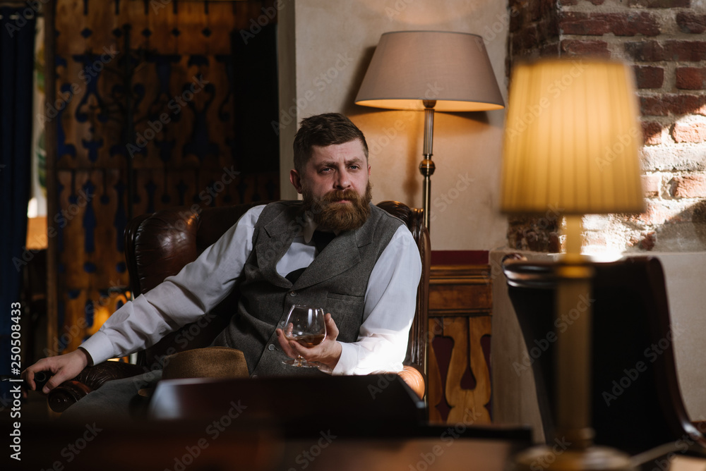 Εκτύπωση καμβά A man in hat wearing vintage suit holding pipe and glass of whiskey sitting on a