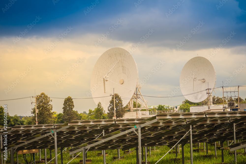 Twin large scale white satellite dish in solar farm under dramatic blue and cloudy sky background.