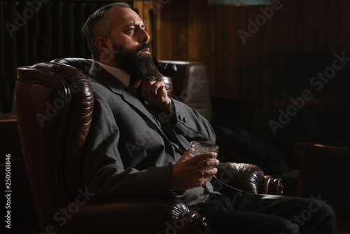 Portrait of serious bearded man with pipe holding glass of whiskey wearing suit and sitting on a big arm chair