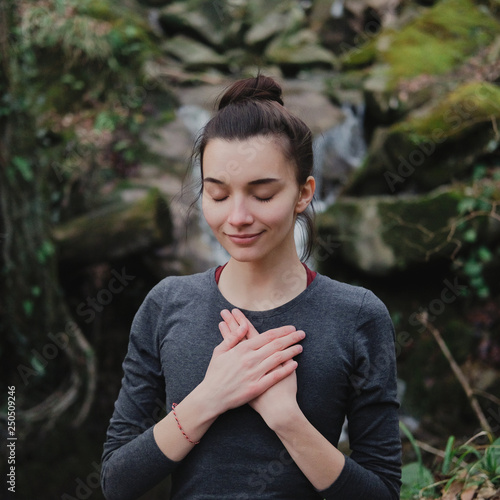 Young woman practicing breathing yoga pranayama outdoors in moss forest on background of waterfall. Unity with nature concept.