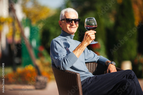 Relaxed old grey-haired man sit at the garden and enjoy glass of wine