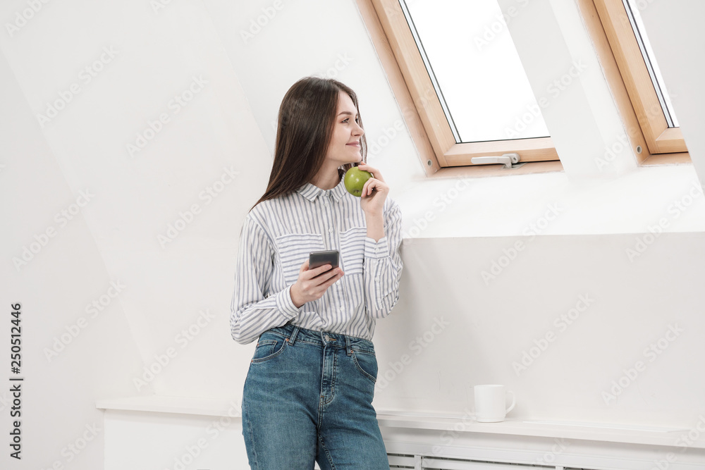 Slender brunette girl with long hair on a white background near the office window. Uses a smartphone and chatting and relaxing during a lunch break. Useful snack green apple. Slender brunette girl with long hair on a white background near the office window. Uses a smartphone and chatting and relaxing during a lunch break. Useful snack green apple.