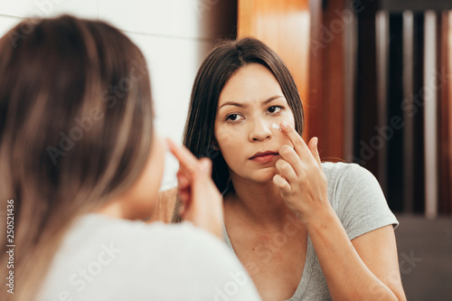 Skin care. Woman applying skin cream on her face in front of mirror