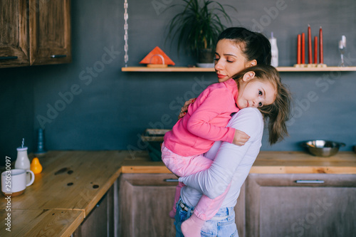 Adorable young woman playing with her little funny daughter in kitchen. Portrait of pretty mother hugging, carrying and watching her litle child. Happy female family indoor lifestyle portrait.