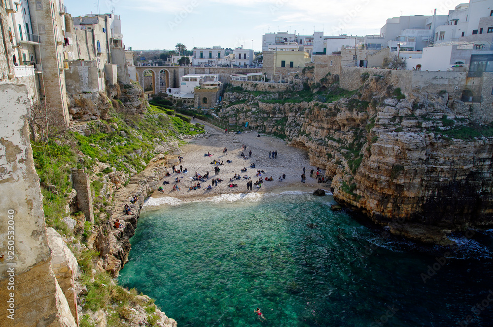 POLIGNANO A MARE, ITALY - MARCH 29th, 2018: People on lovely beach Lama Monachile with dramatic view of cliffs with caves rising from Adriatic sea in Polignano a Mare, Puglia, Italy POLIGNANO A MARE, ITALY - MARCH 29th, 2018: People on lovely beach Lama Monachile with dramatic view of cliffs with caves rising from Adriatic sea in Polignano a Mare, Puglia, Italy
