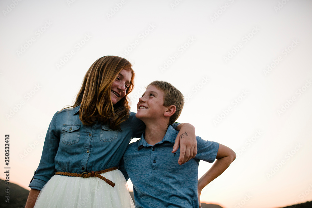 Low angle view of smiling siblings looking each other face to face while standing against clear sky during sunset Low angle view of smiling siblings looking each other face to face while standing against clear sky during sunset