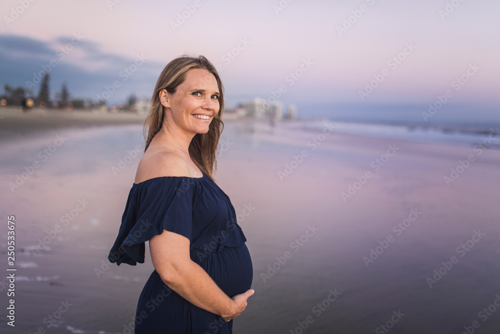 Portrait of smiling pregnant woman with hands on stomach standing at beach against sky during sunset Portrait of smiling pregnant woman with hands on stomach standing at beach against sky during sunset