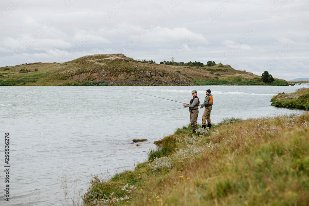 Friends fishing in lake while standing on grassy field against cloudy sky Friends fishing in lake while standing on grassy field against cloudy sky