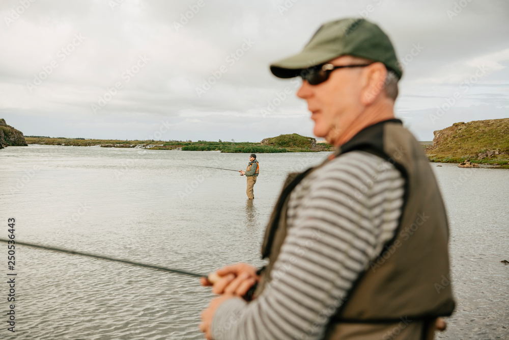 Friends fishing in lake while standing against cloudy sky at Iceland Friends fishing in lake while standing against cloudy sky at Iceland