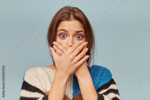 Young woman covering her mouth with hands.Scared girl puts palms on mouth,keeps silence,isolarted on blue background photo