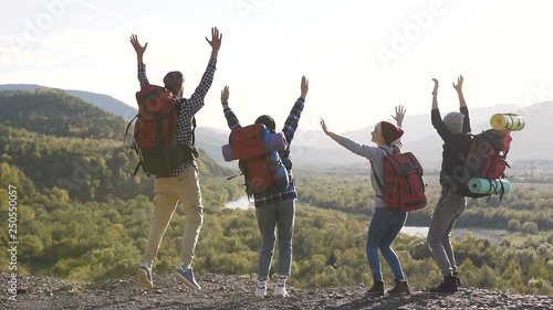 A group of hipsters with tourist backpacks and outfits are bouncing and rejoicing against the backdrop of high mountains and wonderful nature. The concept of success and the conquest of the top. photo