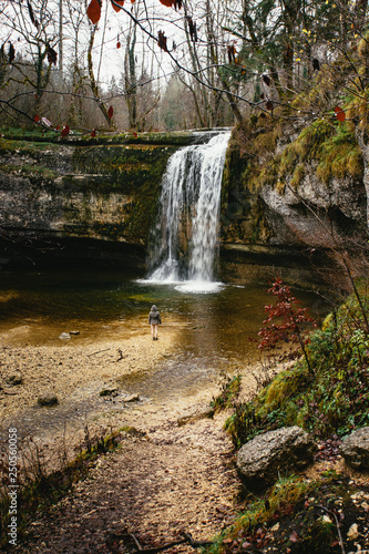 Woman standing in front of a waterfall falling down a roundly shaped stone wall. Cascades du Hérisson, France.