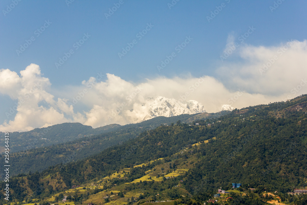 hillside with green forest, fields and houses against the backdrop of the snowy mountain of Annapurna with white clouds and blue sky