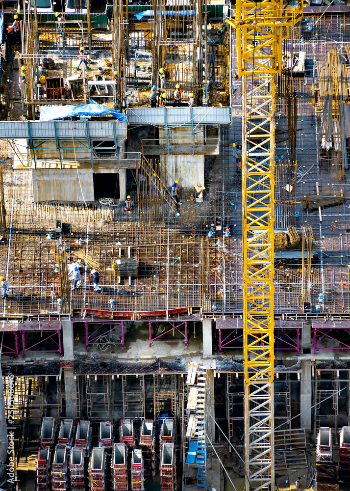Aerial view of a building construction site in progress with tower crane, scaffolding, formwork, construction materials, equipment, and work crews placing reinforcing steel bars.