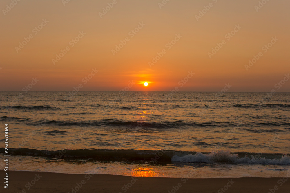 Waves at the sandy shore against the background of the ocean and the evening sunset sky with a sunny path to the sea
