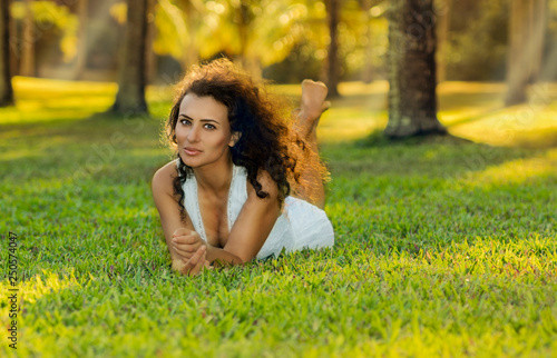 Beautiful curly-haired young woman in beige dress lays on the grass in the garden. Far North Queensland, Thala Beach, Australia. photo