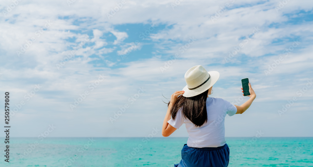 Young Asian woman wear straw hat use smartphone taking selfie at wooden pier. Summer vacation at tropical paradise beach. Happy girl travel on holiday. Woman enjoy and relax life. Summer vibes.