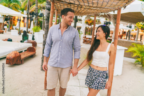 Summer vacation happy couple at tropical beach resort hotel walking to the beach. Young interracial people holding hands in casual clothes. Caribbean or Miami travel holiday lifestyle. © Maridav