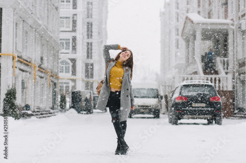Joyful winter young woman chilling in snowfall on strett in big city. Fashionable model, travel with backpack, smiling with closed eyes, enjoying snowing, snowflakes, christmas mood © Look!