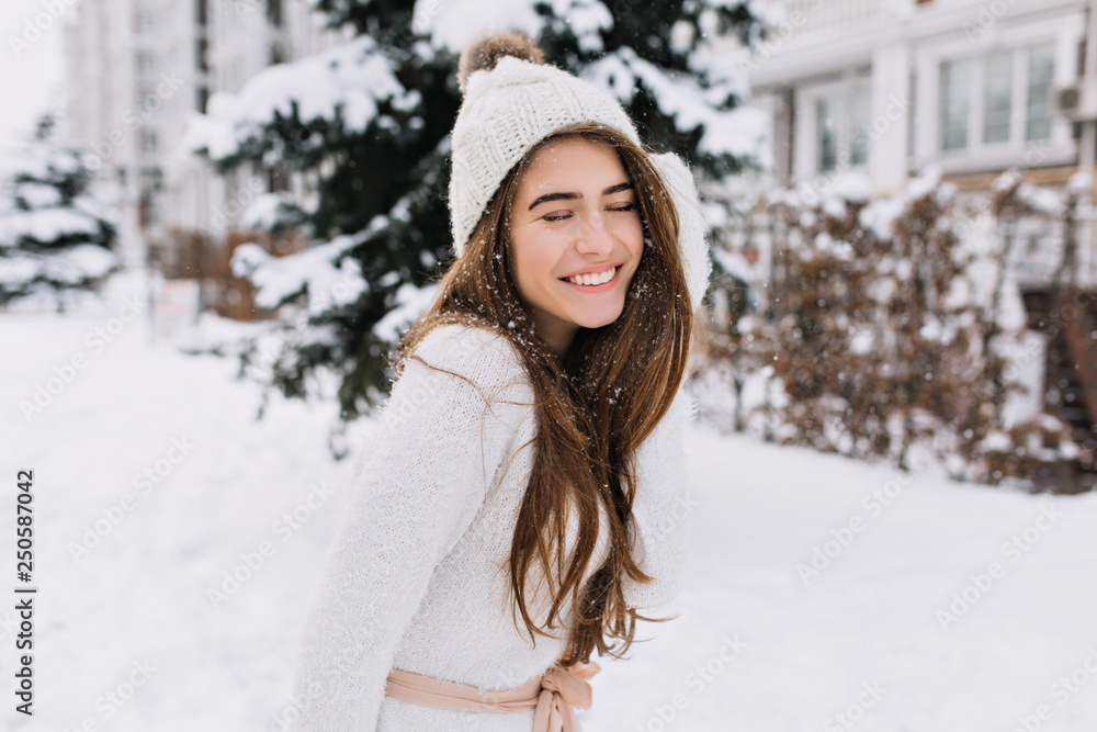 Outdoor portrait of fascinating brunette lady posing with romantic smile on snowy background. Photo of pretty laughing girl in knitted hat and soft sweater on the winter street.. Outdoor portrait of fascinating brunette lady posing with romantic smile on snowy background. Photo of pretty laughing girl in knitted hat and soft sweater on the winter street..