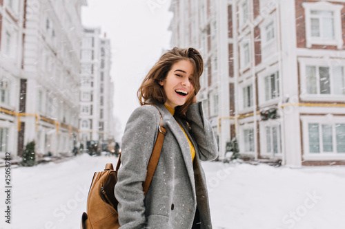 Happy winter time in big city of charming girl walking on street in coat with backpack. Enjoying snowfall, expressing positivity, smiling to camera, joyful cheerful mood, true emotions, new year mood