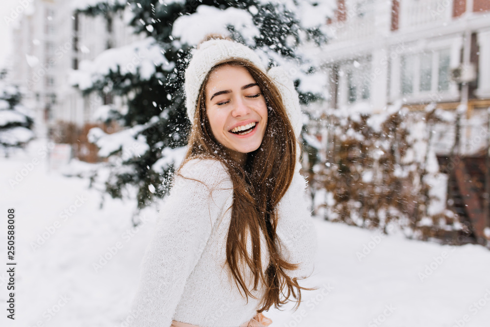 Happy winter moments of joyful young woman with long brunette hair, white winter clothes having fun on street in snowing time. Expressing positivity, true brightful emotions, smiling with closed eyes Happy winter moments of joyful young woman with long brunette hair, white winter clothes having fun on street in snowing time. Expressing positivity, true brightful emotions, smiling with closed eyes