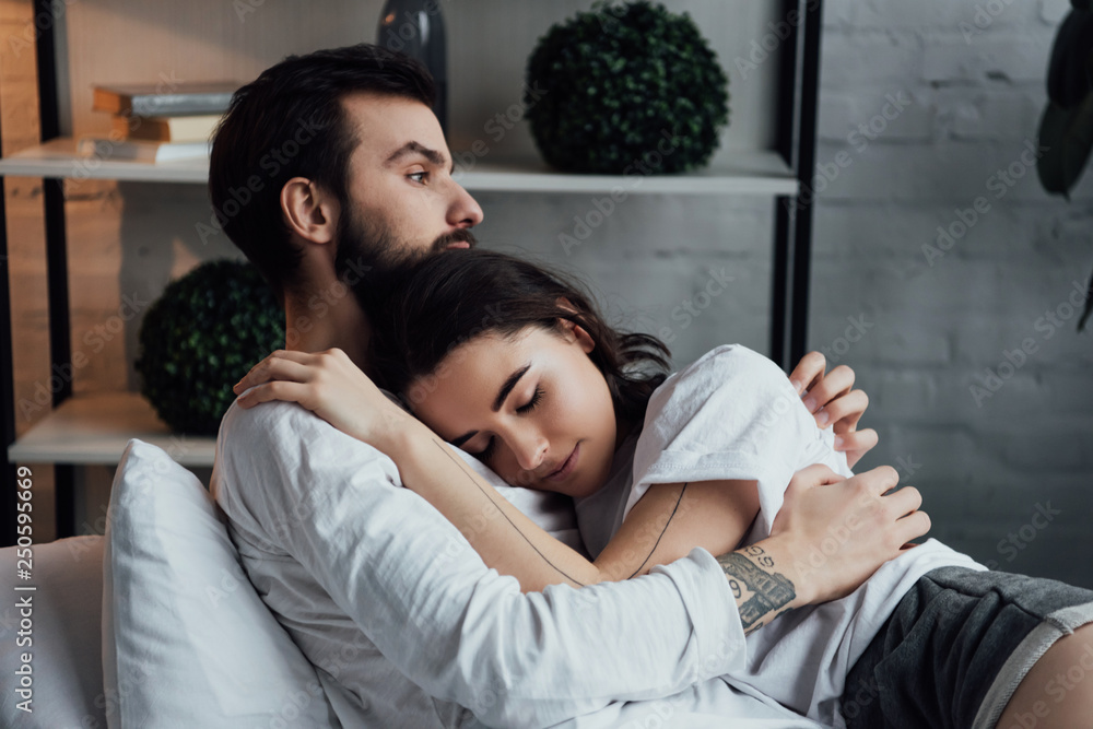 attractive young tattooed couple embracing while lying in bed at home